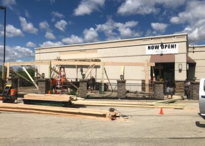 Construction work in progress at the front of a commercial building with a "now open" banner, featuring workers, materials, and machinery.