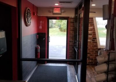 Interior view of a building entrance with an open door, showing a black mat, a fire extinguisher, and a view of the outside lawn.