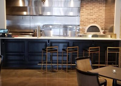 Modern restaurant interior featuring a chandelier, polished concrete floor, and an open kitchen with stainless steel appliances and dark wood accents.