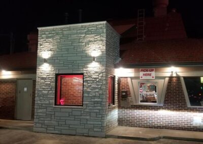 Nighttime view of a fast food restaurant's drive-thru window, illuminated, with a brick facade and a "pick-up here" sign.