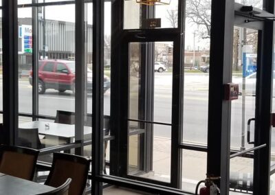 Interior view of a cafe with large glass doors facing a street, featuring a chandelier, tables, and a fire extinguisher near the entrance.