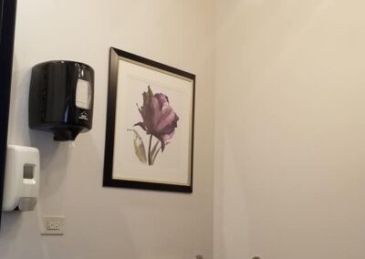 A modern bathroom with black walls, a white ceiling, gold light fixture, framed floral art, and standard white fixtures including a toilet and sink.