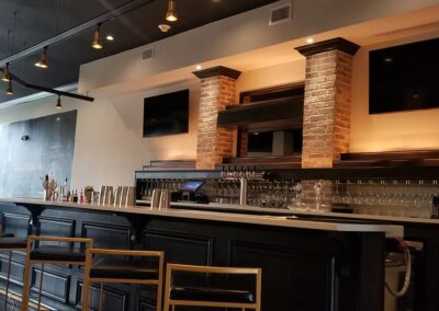 Modern bar interior with high stools, dark countertops, and exposed brick columns, showing an array of glassware and two large televisions.