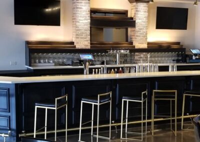 Modern bar interior with black stools, stone pillars, and two large tv screens under a dark ceiling.