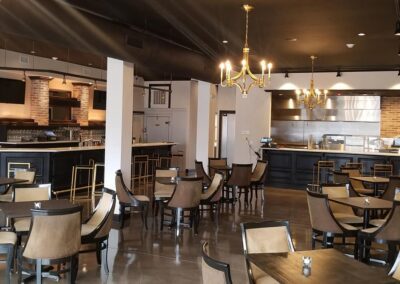 Interior of a modern cafe with black chairs, wooden tables, exposed brick walls, and a chandelier, adjacent to an open kitchen area.