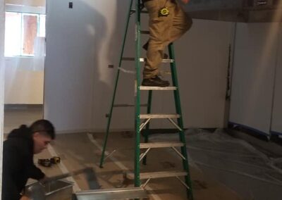 Two workers installing hvac ductwork in a building under construction, one on a ladder and another on the floor.