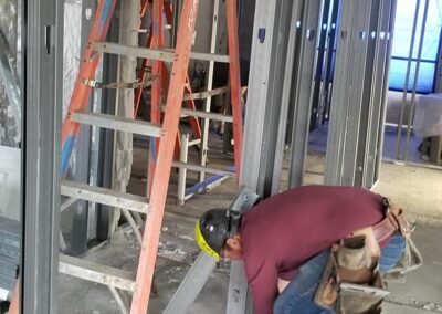 A construction worker uses a drill on a metal stud near an orange ladder inside an unfinished building.