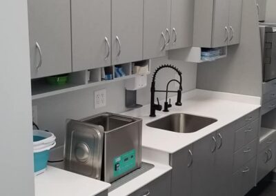 A modern office kitchenette featuring gray cabinets, stainless steel sink, and various kitchen appliances.