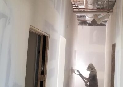 Worker spray painting walls white in a construction site interior, surrounded by exposed insulation ducts and electrical wiring.