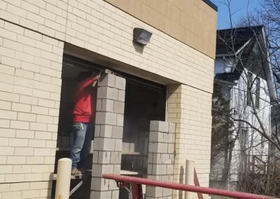 A worker in a red shirt demolishes a brick wall of a building, debris scattered around on the ground.