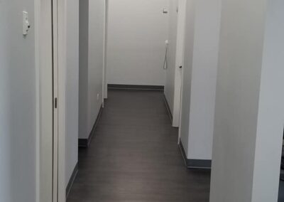 A narrow office corridor with closed doors on either side, gray flooring, white walls, and an exit sign in the distance.