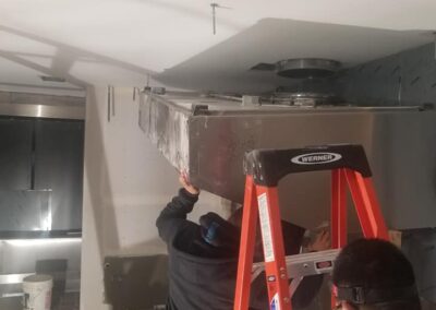 Two workers installing a large air duct in a commercial kitchen, standing on ladders and using tools.