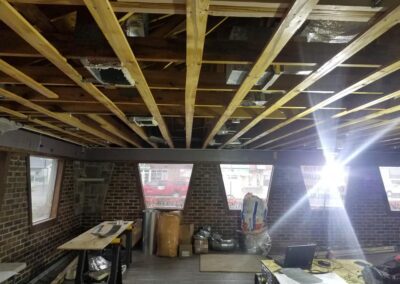 Interior construction space with exposed wooden beams, brick walls, and materials scattered on the floor, sunlight streaming in through a large window.