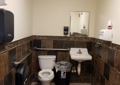 Public restroom interior with a toilet, sink, and mirror, featuring dark brown tiled walls and a wooden floor.