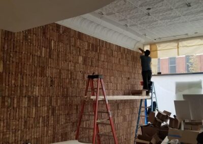 A person on a ladder installs tiles on a large, curved wall inside a room under construction.