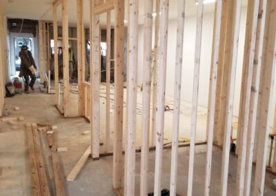 Interior construction site with wooden framing for rooms, and a worker visible in the background. tools and building materials are scattered around the floor.