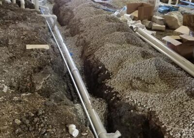 Trenches dug inside a building under construction for plumbing installation, with exposed pipelines and debris around. bright light streams in from an open doorway.