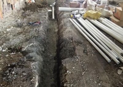 Trenches dug inside a building under renovation with exposed pipes and construction debris.