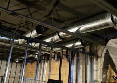 A construction worker installs ductwork in a building's ceiling framed with metal studs, with a ladder nearby.