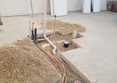 Concrete floor of a room under construction with exposed plumbing and electrical wires, surrounded by gravel and some construction tools.