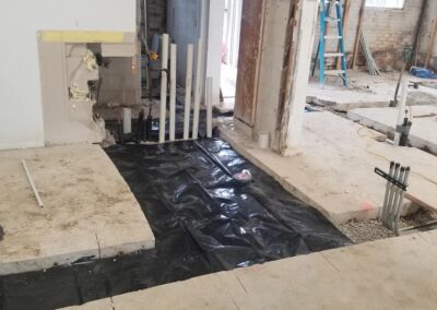 Interior of a building under construction with exposed pipes and a plastic-covered floor in a room with unfinished walls.