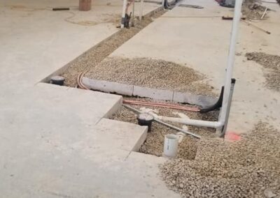 An indoor construction site showing exposed pipes and cables running through a trench in a concrete floor, surrounded by tools and materials.