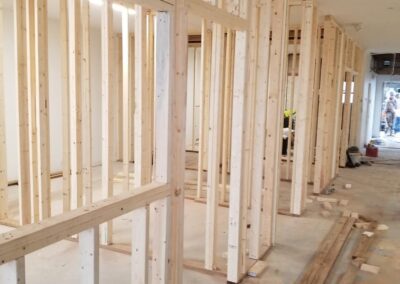 Interior view of a building under construction with exposed wooden stud walls and a construction worker visible in the background.