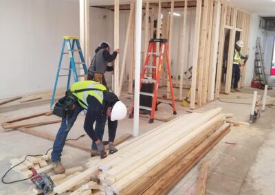 Two construction workers frame a wall inside a building, surrounded by wooden planks and ladders.