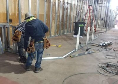 Two construction workers installing ductwork in an unfinished building interior with visible metal studs and insulation.