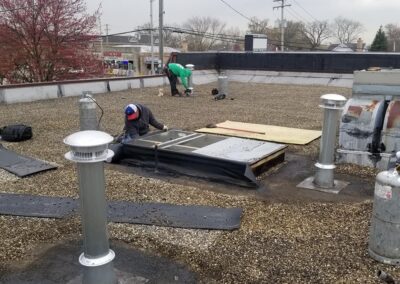 Workers repair a flat roof with gravel, using tools near various roof vents and pipes.