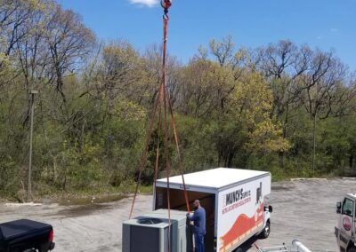 A crane lifts an hvac unit onto a truck in a parking lot surrounded by trees. a worker oversees the operation.