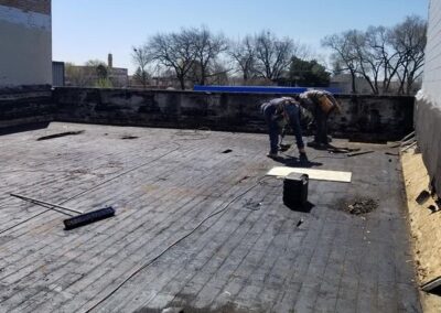 Two workers repairing a flat roof with tools and materials around them, surrounded by brick walls under a clear sky.