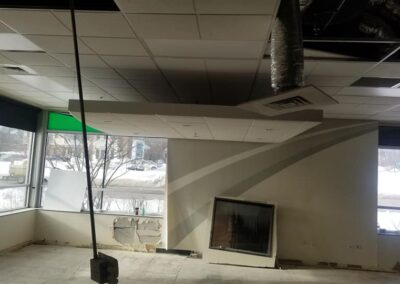 Interior of a building under renovation featuring exposed ductwork, partially removed ceiling panels, and large windows overlooking a snowy scene outside.