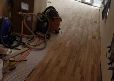 A man kneels while working on new flooring installation in a room cluttered with construction tools and materials.