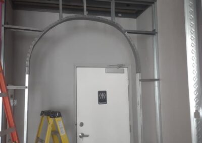 A white door with a number plaque under an arched metal frame in a construction site, with exposed wiring and a ladder to the side.