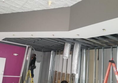 A worker on a ladder installs ductwork in a room with purple walls and a partially completed drop ceiling.