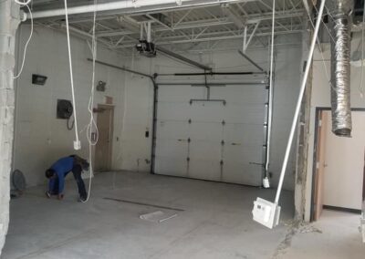 A worker kneels near a garage door in an under-construction area, surrounded by exposed wires and ductwork.