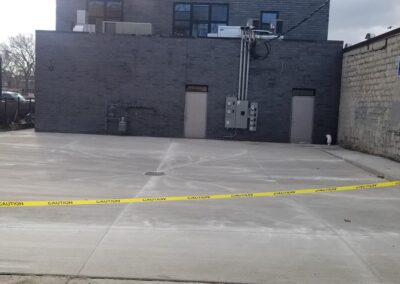 An empty parking lot cordoned off with yellow caution tape, featuring a grey building and a partial view of a black vehicle on the right.