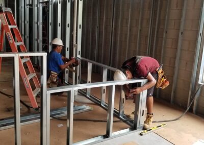 Two construction workers install metal frames inside a building, one using a handheld tool, with a ladder nearby.