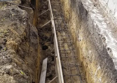 Worker walking beside a trench with reinforced steel bars and wooden planks, preparing for construction beside a concrete wall.