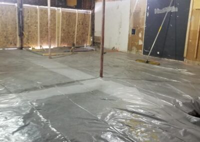 An unfinished interior space with exposed walls, a concrete floor partially covered by a plastic sheet, and construction materials.