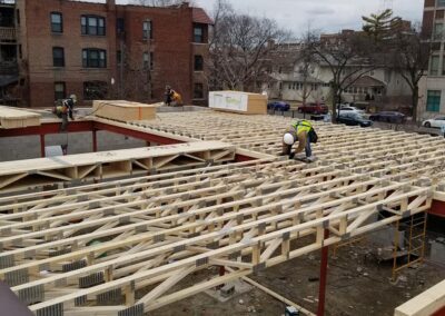 Construction workers install wooden trusses on a building structure, with residential buildings and a cloudy sky in the background.