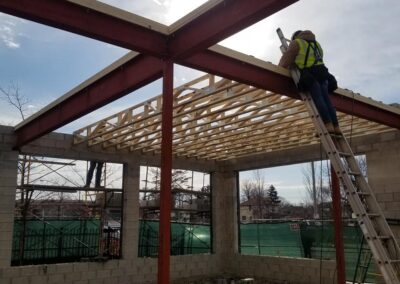 Construction worker on a ladder welding a metal beam on a building frame with wooden trusses under a clear sky.