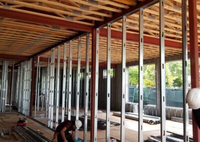 Interior of a construction site with metal stud framing and a wooden roof, featuring a worker bending over near materials.