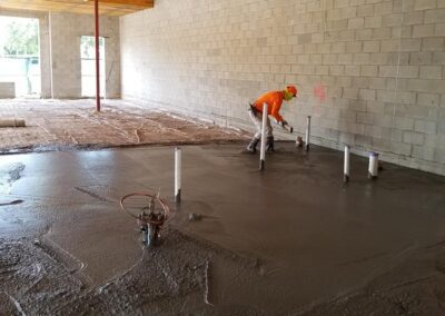 Worker in orange shirt smoothing wet concrete inside a building under construction with exposed beams and block walls.