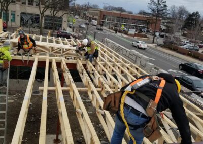 Construction workers in safety gear assemble a wooden framework on a building site, with a city street visible in the background.