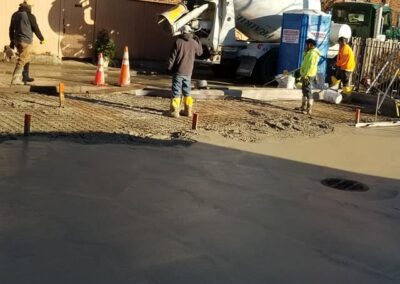 Construction workers pour concrete from a mixer truck into a foundation at a building site, with safety cones and equipment visible.