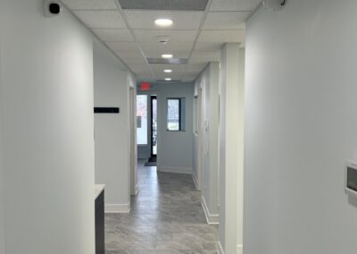 Narrow hallway in a modern building with gray tiled floors, white walls, and evenly spaced doors, illuminated by overhead lighting.