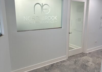 Modern orthodontic office lobby with a frosted glass panel showcasing the clinic's logo and name, next to a glass door.